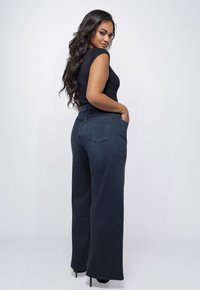 Vrouw met lang donker haar, gekleed in een zwarte mouwloze top, donkere wijde jeans en hakken, staand in een zijaanzicht met haar handen in de zakken.