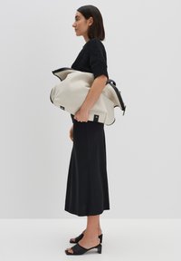 Große, cremefarbene Canvas-Tasche mit schwarzen Lederakzenten, Reißverschluss und silbernen Beschlägen, gehalten von einer Person, die ein schwarzes gestricktes Oberteil und einen langen schwarzen Rock trägt.