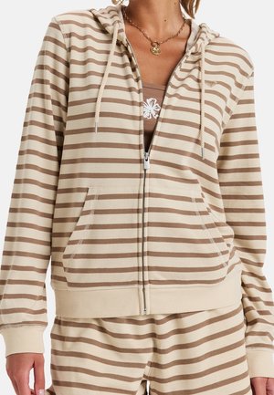Kobieta ubrana w beżowo-brązową bluzę na zamek z kapturem i przednimi kieszeniami oraz pasujące spodnie w paski, ozdobiona złotym łańcuszkiem.