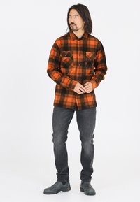 Orange og sort rutet flannel skjorte med to brystlommer, knaplukning, lange ærmer, båret over mørkegrå jeans og ankelstøvler.