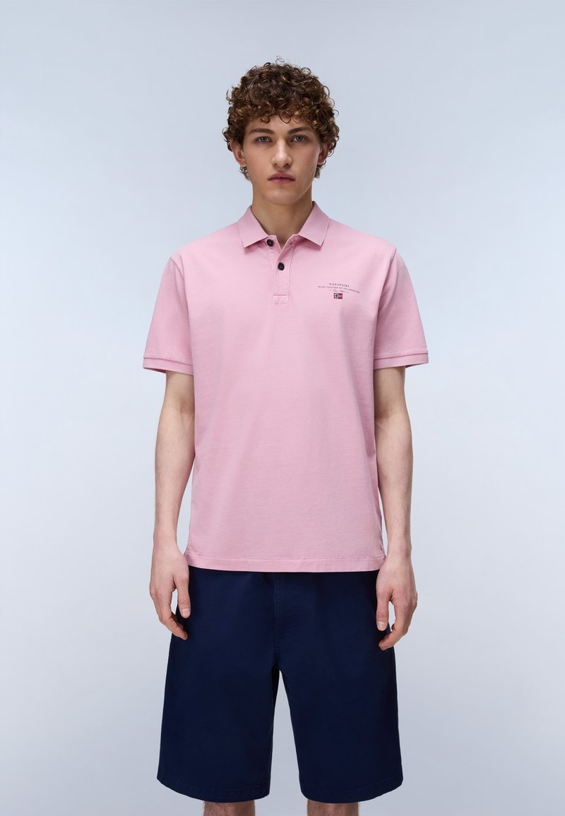 Jeune homme aux cheveux bouclés portant un polo rose clair et un short bleu foncé, debout devant un fond clair uni.