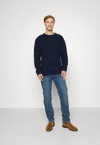 Navy geribde trui met lange mouwen, ronde hals; gecombineerd met blauwe spijkerbroeken en bruine suède enkellaarsjes. Model staat voor een neutrale achtergrond.