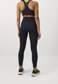 Černá sportovní podprsenka s racerback střihem a tmavě modré legíny s vysokým pasem. Černé sportovní boty s neonově zelenými akcenty. Hladká, elastická tkanina.