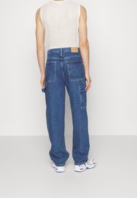 Pantalones cargo de mezclilla con corte holgado, dos grandes bolsillos laterales y un parche de cuero marrón en la parte trasera. Llevados con una blusa sin mangas blanca.