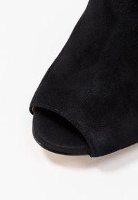 Musta nubukista valmistettu avonainen kenkä, jossa on sileä pinta, yksinkertainen muotoilu ja kaarevat reunat. Pohja on litteä ja hieman teksturoitu.