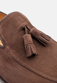 Moccasini in suede marrone con due nappine decorative, texture liscia, punta arrotondata e cuciture visibili lungo i bordi.