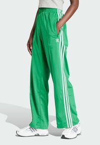 Groene sportbroek met glanzende afwerking, voorzien van witte zijstrepen en een elastische tailleband. Inclusief zakken en een Adidas logo.