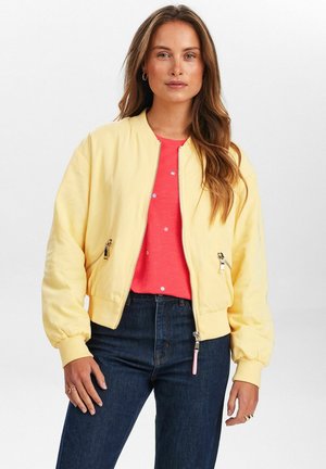 Gelbe Bomberjacke mit gerippten Bündchen und Saum, zwei Reißverschlusstaschen und einem glatten Stofffinish, getragen über einem korallenfarbenen Hemd und dunklen Jeans.