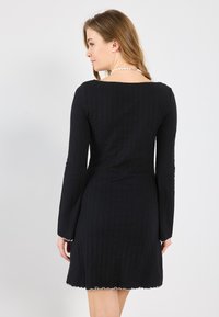 Abito nero a maniche lunghe con texture a coste, caratterizzato da una scollatura quadrata sulla schiena e dettaglio con orlo ondulato bianco.
