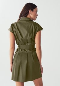 Vestito camicia verde oliva con maniche corte a cappuccio, vita cintrata e gonna plissettata. Tessuto liscio con design strutturato e colletto.