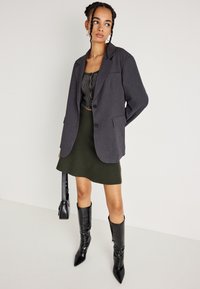 Žena s opletenými vlasmi, oblečená v prehnane veľkom šedom saku, pruhovanom crop tope, olivovej sukni, čiernych vysokých čižmách a drží čiernu kabelku.