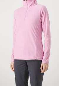 Ljust rosa fleecetröja med halvdragkedja, långa ärmar och en figurnära passform, matchad med mörka byxor för kontrast.