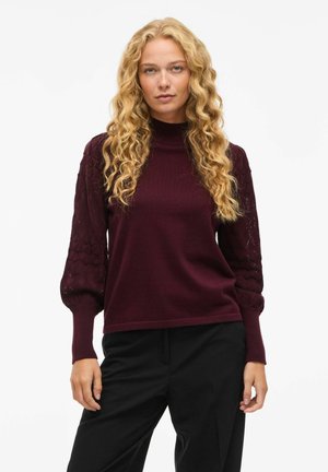 Pull bourgogne à col montant, texturé, avec des manches bouffantes décorées d'un motif en losanges et des poignets côtelés. Associé à un pantalon noir.