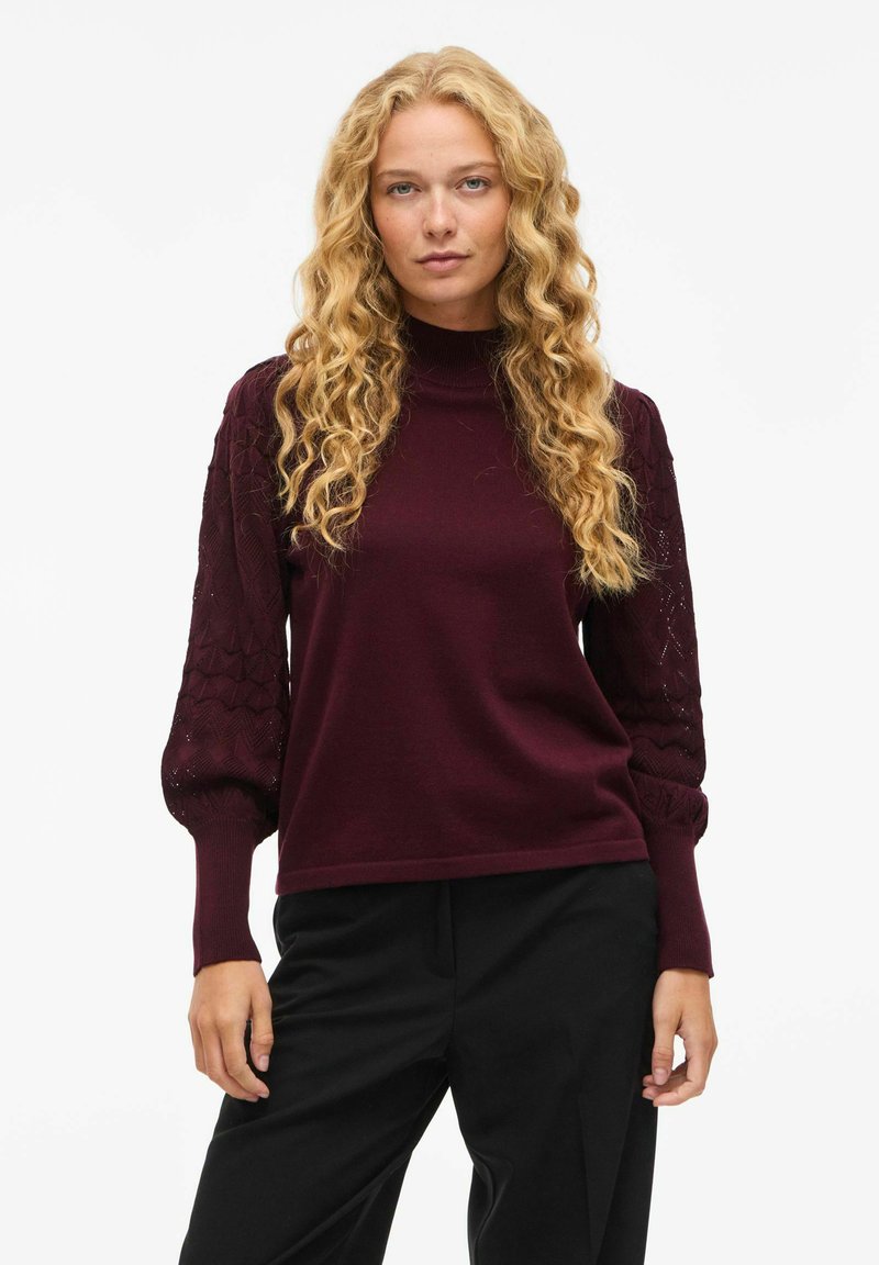Burgundy tröja med hög krage, texturerade, puffiga ärmar med ett diamantmönster och ribbade muddar. Kombinerad med svarta byxor.