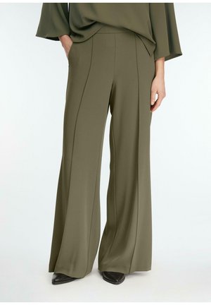 Kobieta w oliwkowozielonych spodniach o szerokich nogawkach i pasującym topie, stojąca z jedną ręką w kieszeni, w czarnych spiczastych butach.