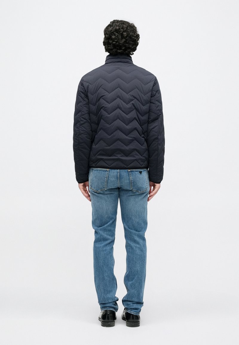 Veste matelassée noire avec un col montant, présentant un motif en chevron. Portée avec un jean bleu et des chaussures noires, vue de dos.