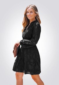 Vestido negro con estampado floral, diseño en capas, manga larga y escote en V. Combinado con un bolso de mano marrón, sobre un fondo claro.