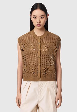 Kvinde med glat mørkt hår iført ærmeløs brun strikket vest med knapper foran og cremefarvede plisserede bukser mod ensfarvet baggrund.