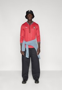 Κόκκινη μακριά μπλούζα με κυματιστό μοτίβο και μπλε λεπτομέρειες, σε συνδυασμό με χαλαρό μαύρο παντελόνι και ανοιχτό μπλε πουλόβερ δεμένο στη μέση. Μαύρο καπέλο τύπου bucket.