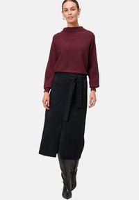 Woman wearing a burgundy ribbed sweater, black midi skirt with a front slit and belt, and black knee-high boots, standing against a plain background.