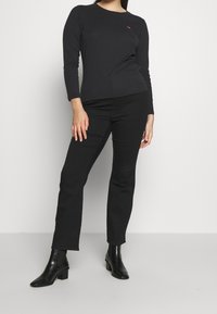 Schwarzes Langarmoberteil mit einem runden Halsausschnitt und einem kleinen roten Logo, kombiniert mit schwarzen Slim-Fit-Hosen und Ankle Boots. Glatte Textur insgesamt.