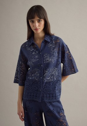 Frau trägt marineblaue Spitzen-Bluse mit Knöpfen und passende Hose, steht vor neutralem Hintergrund.