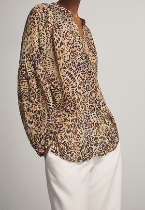Femme portant un chemisier ample à manches longues avec un imprimé abstrait marron et beige, associé à un pantalon blanc, les mains dans les poches.