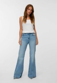 Jonge vrouw in een witte tanktop en lichtblauwe flared jeans die tegen een effen lichte achtergrond staat.
