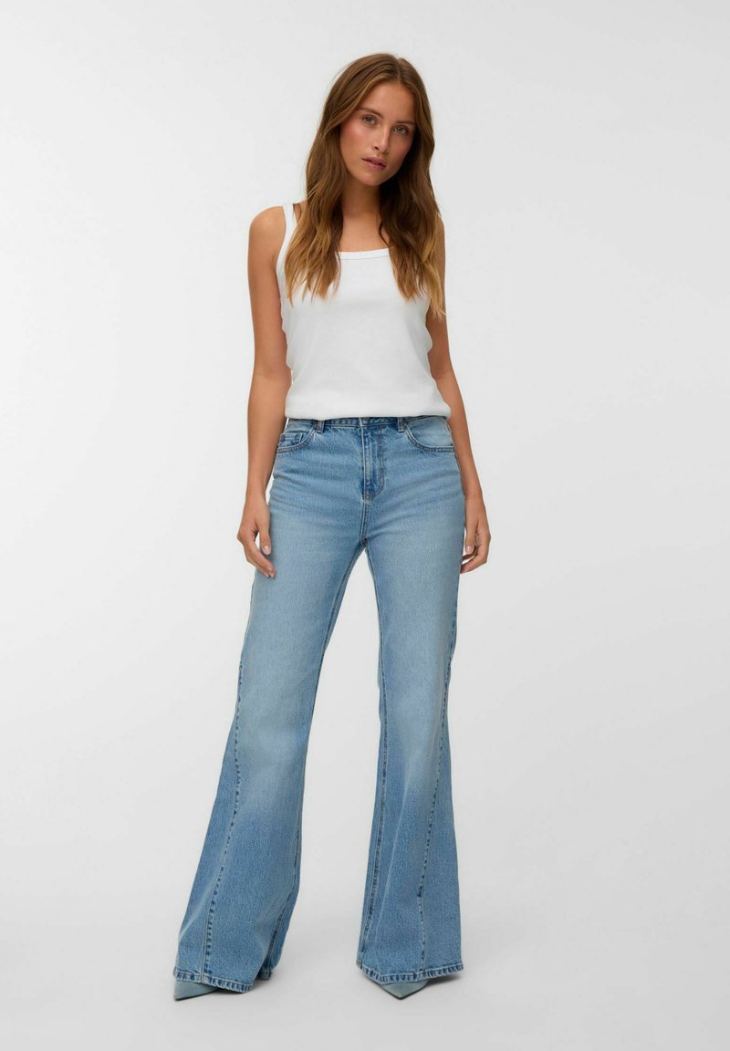 Jonge vrouw in een witte tanktop en lichtblauwe flared jeans die tegen een effen lichte achtergrond staat.
