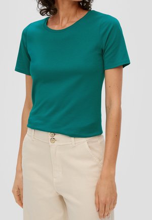 Kobieta nosząca dopasowany turkusowy t-shirt z krótkim rękawem wpuszczony w beżowe, wysokie spodnie z podwójnym zapięciem na guziki.