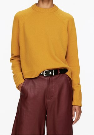Personne portant un pull jaune moutarde rentré dans un pantalon en cuir bordeaux taille haute avec une ceinture noire et une boucle en argent. Mains dans les poches.