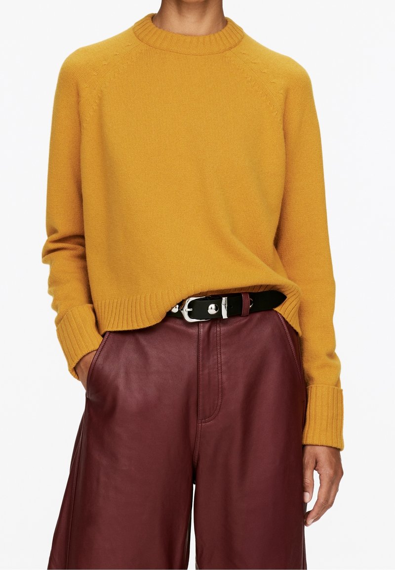 Personne portant un pull jaune moutarde rentré dans un pantalon en cuir bordeaux taille haute avec une ceinture noire et une boucle en argent. Mains dans les poches.