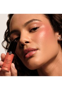 Giorgio Armani Luminous Silk Cheek Tint, korall-oransje væske i en glastype med svart lokk, holdt av en person mot en nøytral bakgrunn.