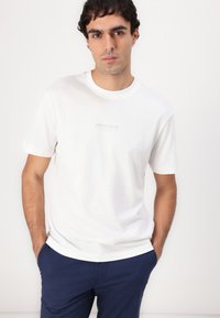 Hvid t-shirt med korte ærmer lavet af bomuld, med et minimalistisk logo trykt i grå på brystet. Casual pasform med rund halsudskæring.