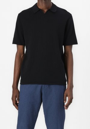 Person trägt ein schlichtes schwarzes kurzärmeliges Poloshirt und locker sitzende blaue Hosen, steht vor weißem Hintergrund.