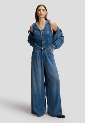 Frau trägt weite Jeanshosen und eine passende Jeansweste über einer Jeansjacke, steht mit den Händen in den Taschen vor einem einfarbigen Hintergrund.