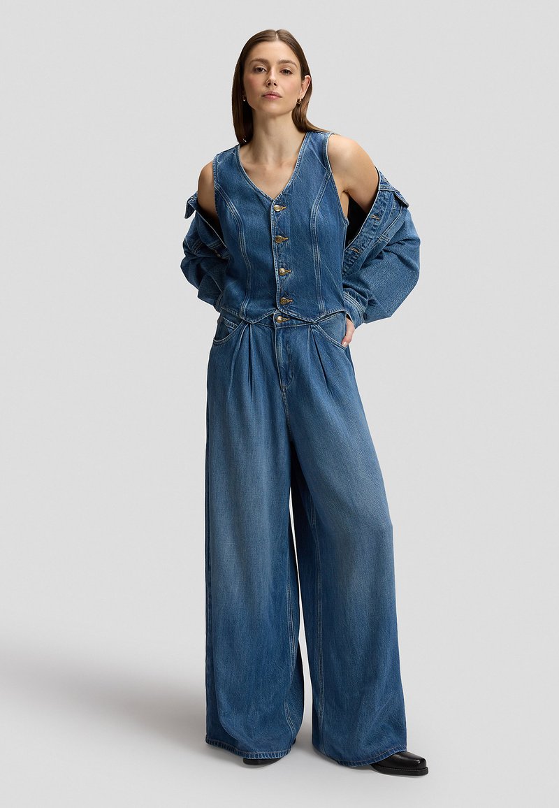Femme portant un pantalon en jean à jambes larges et un gilet en jean assorti par-dessus une veste en jean, debout les mains dans les poches devant un fond uni.