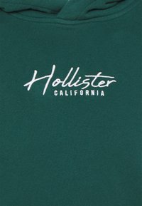 Sudadera con capucha verde hecha de algodón, con capucha ajustable mediante cordón y logotipo blanco "Hollister California" en cursiva en el pecho.