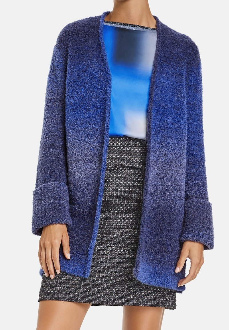 Femme portant un cardigan doux bleu ombré, un haut à imprimé abstrait bleu et une mini-jupe tissée noire et blanche, debout devant un fond uni.