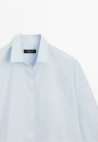 Světle modrá košile s klasickým límečkem a náprsní kapsou, elegantně vystavená na bílém pozadí.