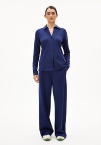 Marineblauwe longsleeve overhemd met kraag en voorzak, gecombineerd met marineblauwe wijde broek en witte sneakers met groene accenten.