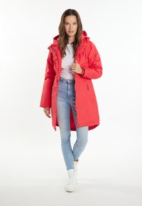 Bright red parka coat with a hood, featuring a front zipper, snap buttons, and side pockets, paired with light blue jeans and white sneakers.