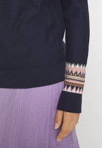 Pull marine avec des poignets géométriques à motifs roses, blancs et noirs, porté avec une jupe plissée lavande, montrant une main reposant le long du corps.