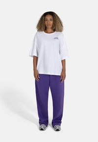 White oversized t-shirt with short sleeves, featuring purple logo. Paired with loose purple pants and silver sneakers, set against a plain background.