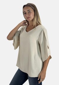 Blusa beige a V con maniche corte larghe e tessuto testurizzato. Abbinata a jeans scuri, la blusa ha una vestibilità comoda e un drappeggio sottile.