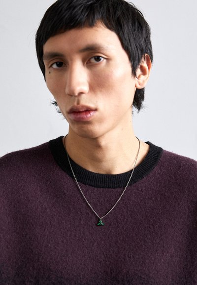 Maglione viola con scollatura a coste nera, indossato con una catena d'argento che presenta un piccolo ciondolo verde. Tessuto morbido, design aderente.