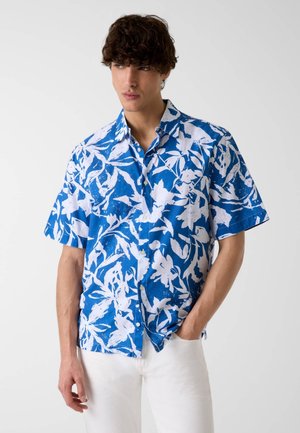 Jeune homme portant une chemise à manches courtes fleurie bleu et blanc boutonnée et un pantalon blanc, debout avec une main dans la poche.