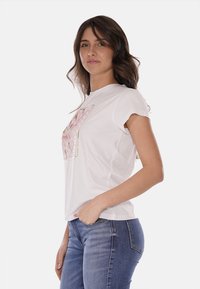 T-shirt bianco a maniche corte con collo rotondo, caratterizzato da una stampa floreale sulla parte anteriore e dettagli decorativi a forma rettangolare.