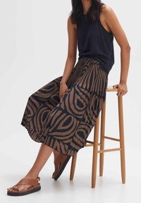 Kvinna som sitter på en träpall iklädd en svart ärmlös topp, ett brun och svart mönstrat midi-skjol, och bruna sandaler.