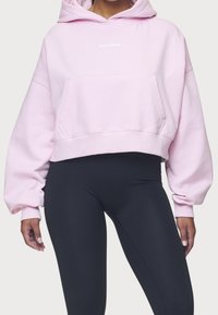 Cropped pink hoodie with a front pocket and logo, featuring wide sleeves and a soft texture. Paired with black leggings.